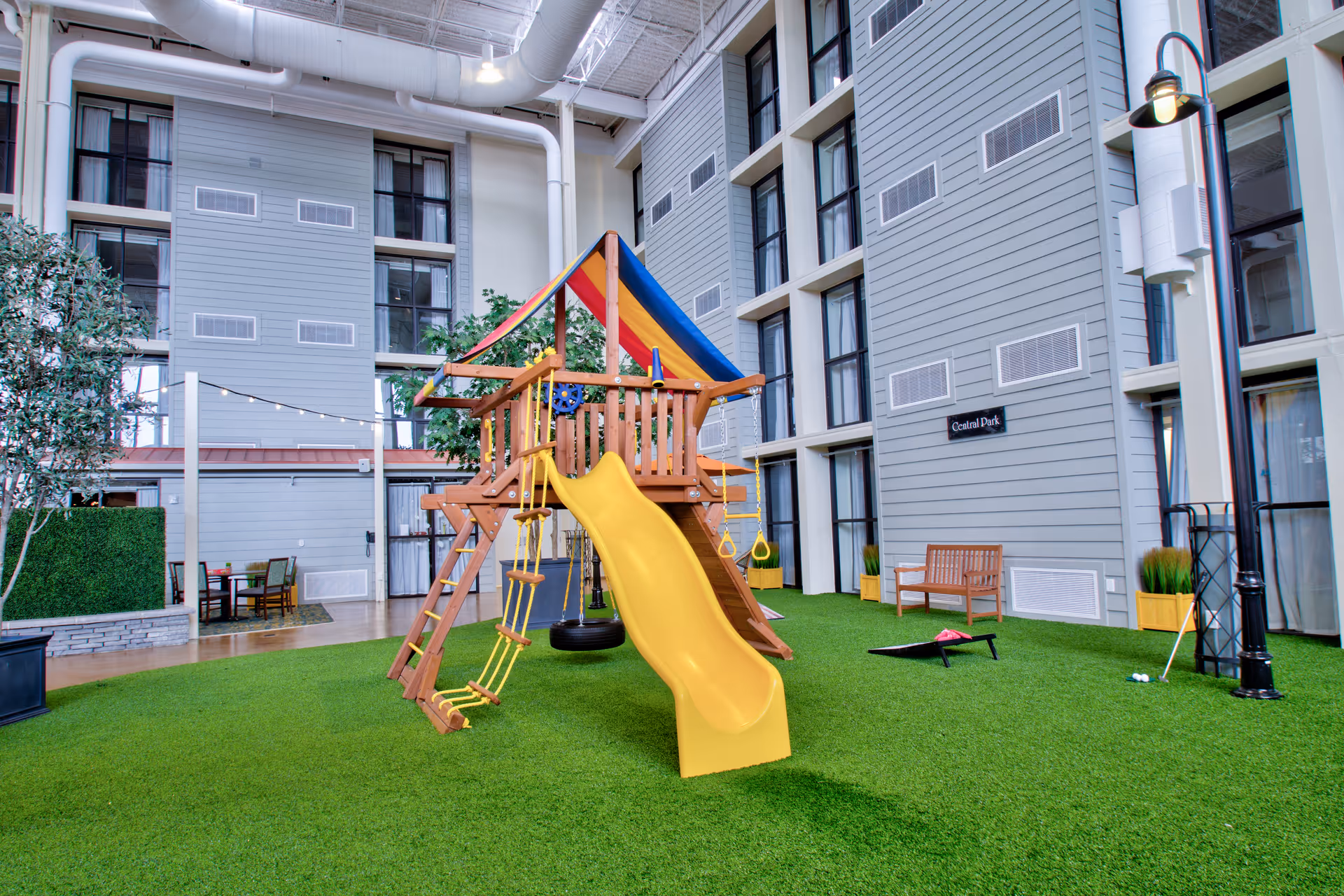 Indoor play area with a wooden playset featuring a yellow slide, climbing ropes, and swings on artificial green grass. Surrounding the play area are tall windows and gray walls with multiple floors visible. There is a wooden bench, a cornhole game, and some potted plants near the walls.