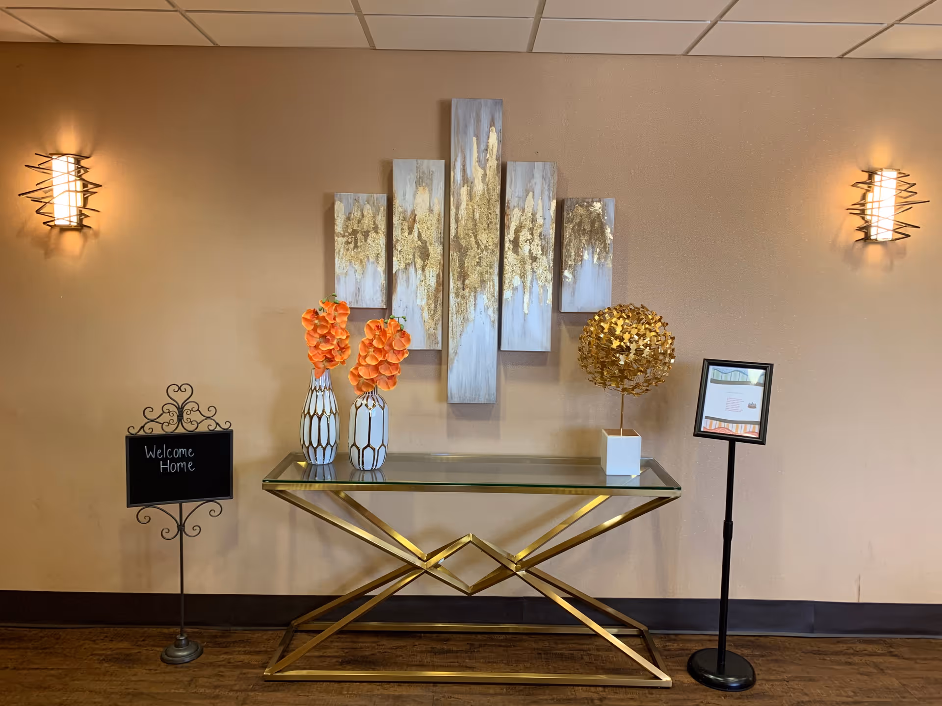 A decorative interior wall with a modern glass table featuring a gold geometric base. On the table are two white vases with orange flowers and a gold spherical sculpture. Above the table is a multi-panel abstract painting with gold and gray tones. On the left side, a small blackboard sign reads 'Welcome Home'. Two wall sconces with warm lighting flank the scene.