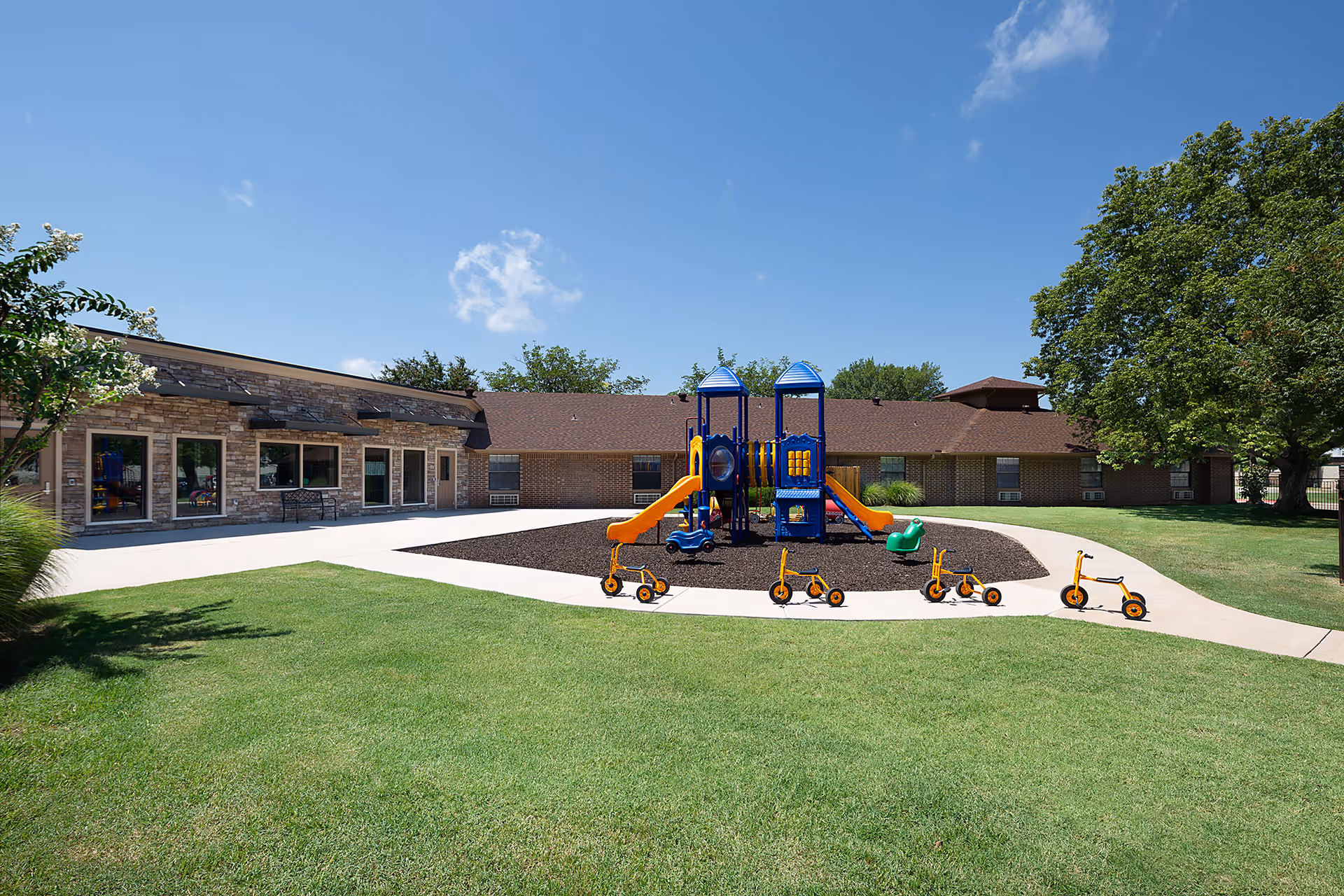 Outdoor playground area with a blue and yellow play structure featuring slides, surrounded by a paved walkway and green grass. Several small orange tricycles are lined up on the walkway. The background shows a single-story brick building under a clear blue sky with some trees.