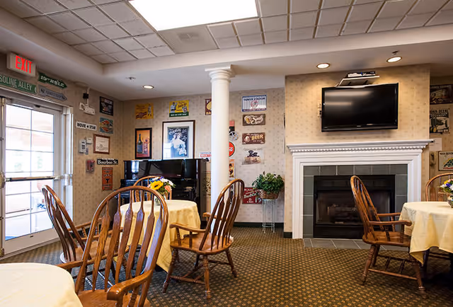 A cozy common area with round tables covered in yellow tablecloths and wooden chairs. There is a fireplace with a mounted flat-screen TV above it. The walls are decorated with various vintage-style signs and posters. A black piano is positioned near a white column, and a glass door with an exit sign is visible on the left side.