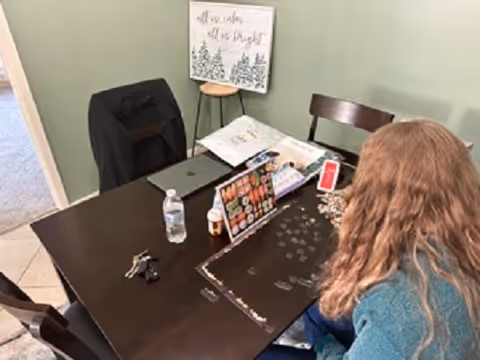 A person with long hair sitting at a dark wooden table working on a puzzle. The table has a water bottle, keys, a laptop, a bottle of pills, and a sign that says 'Welcome'. In the background, there is a green wall with a framed sign that reads 'All we color all we delight' and a small stool.