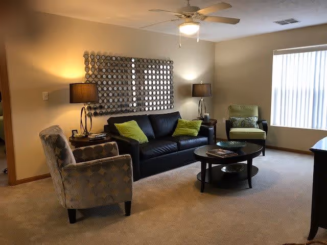 A cozy living room with a black leather sofa adorned with green pillows, two patterned armchairs, a round black coffee table with books and a decorative bowl, two side tables each with a lamp, a ceiling fan with a light, and a window with vertical blinds letting in natural light.