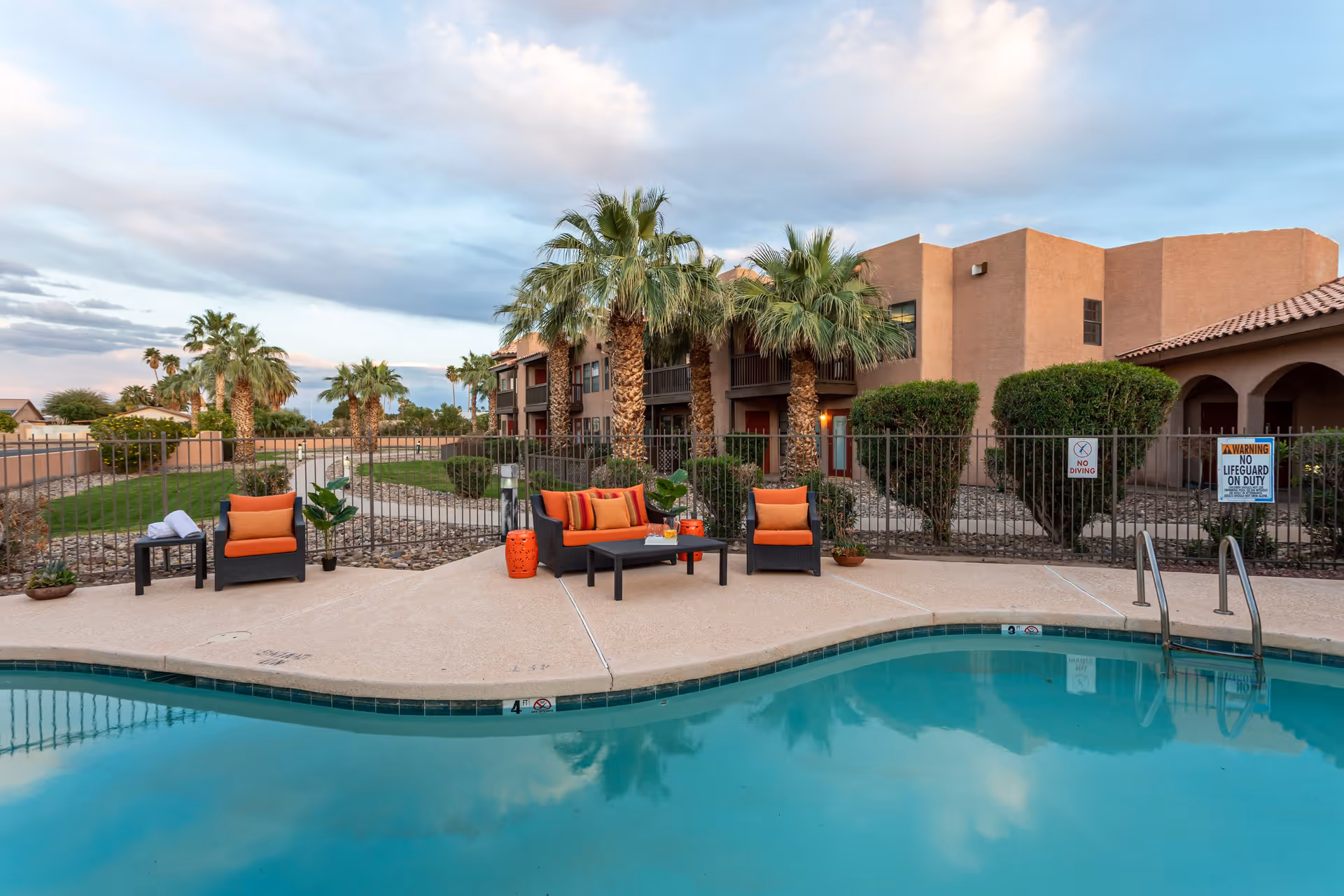 Outdoor swimming pool area at Emerald Springs Senior Living with patio furniture including chairs and a sofa with orange cushions, surrounded by palm trees and a fenced garden area, under a partly cloudy sky.