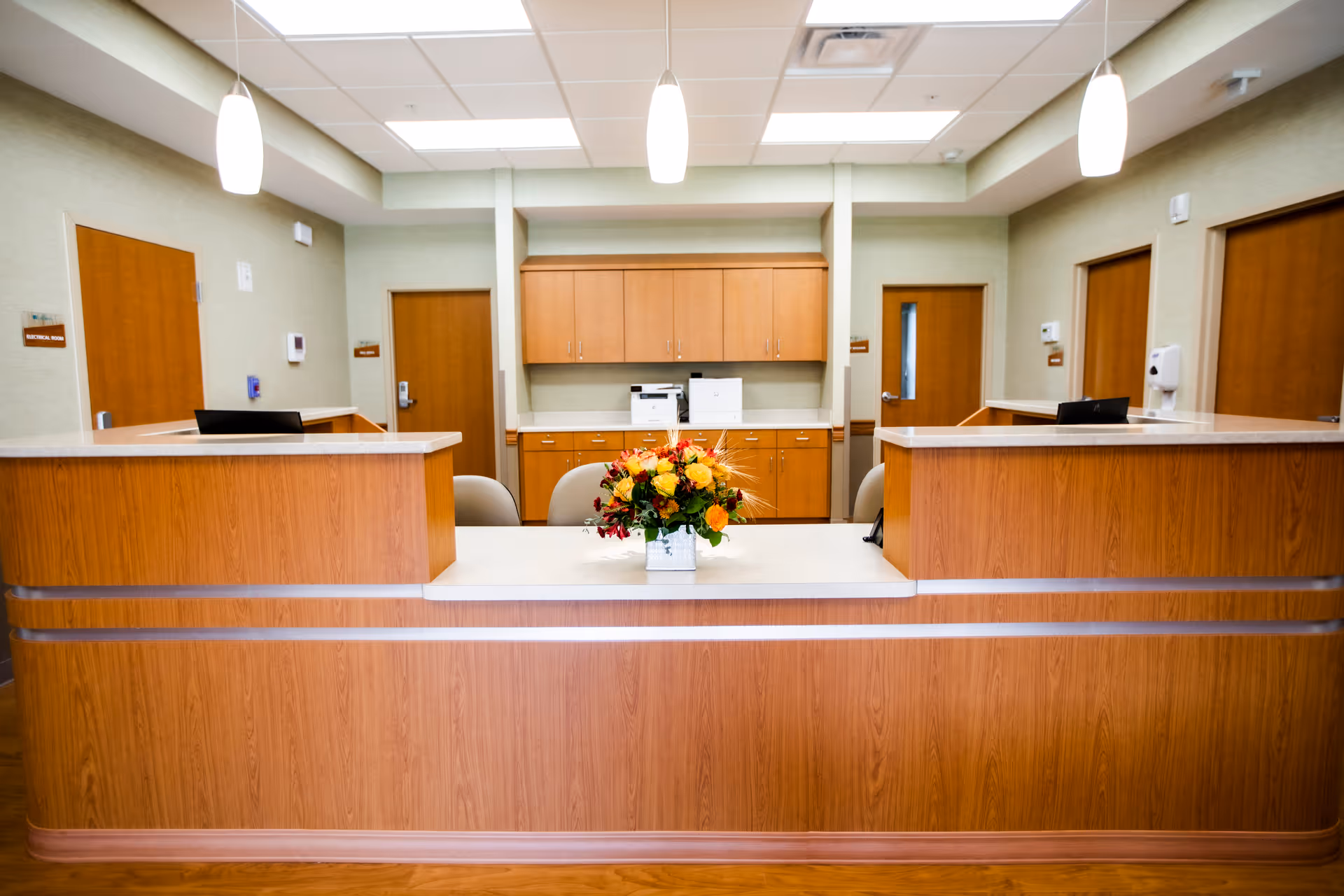Reception desk area in a senior living facility with a wooden counter, a flower arrangement in the center, three pendant lights hanging from the ceiling, and multiple wooden doors and cabinets in the background.