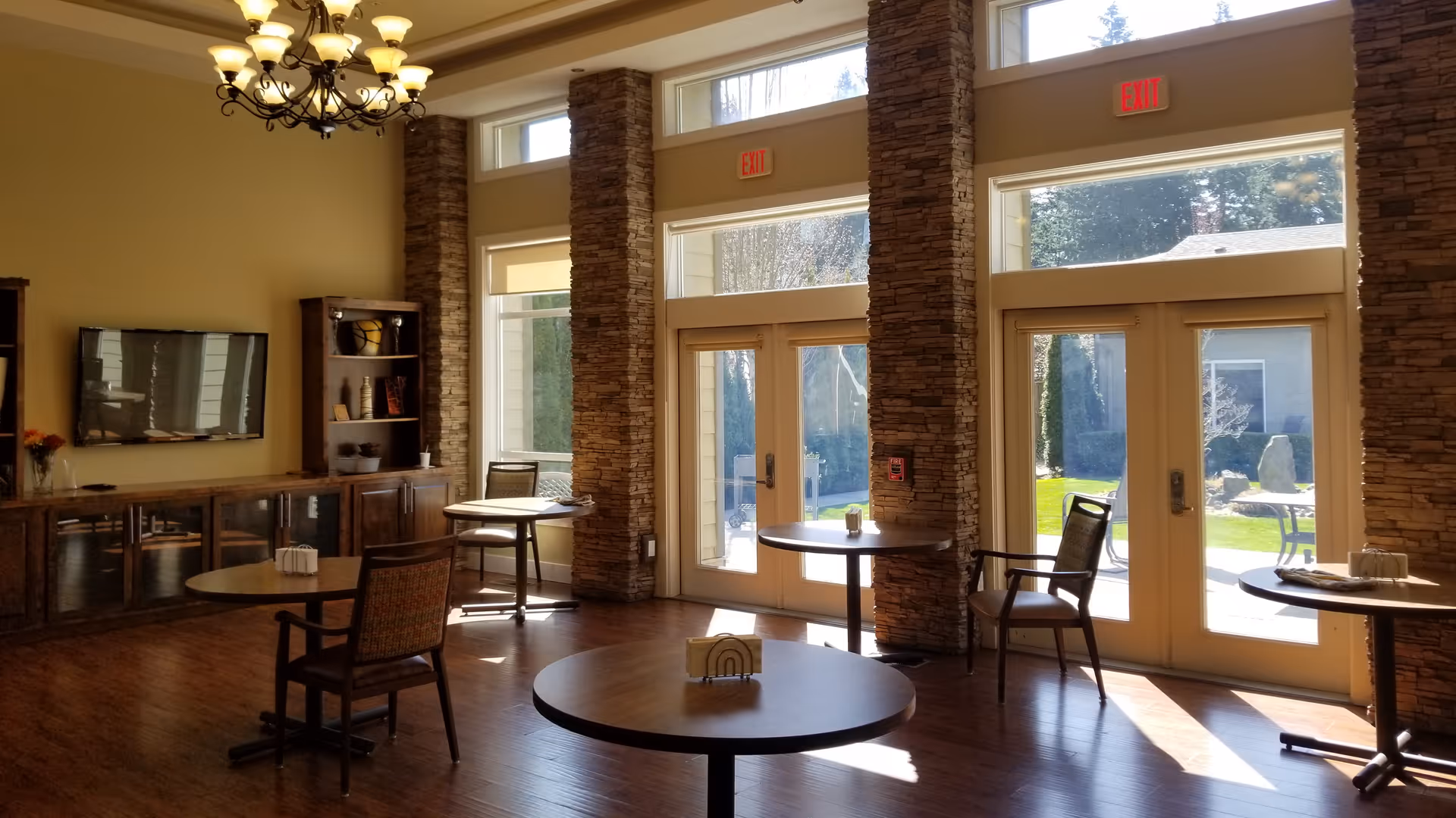 A bright and spacious common area with large windows and glass doors letting in natural light. The room features round wooden tables with chairs, a wooden cabinet with decorative items, a wall-mounted flat-screen TV, and stone pillars between the windows. Outside, a green lawn and patio furniture are visible.