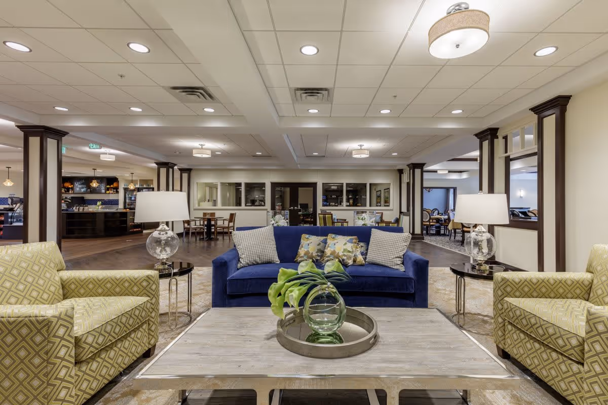 A spacious and well-lit living room area in a senior living facility featuring a blue sofa with patterned cushions, two yellow patterned armchairs, a wooden coffee table with a decorative glass vase and green leaves, and two glass table lamps on either side of the sofa. In the background, there are dining tables and chairs, with a kitchen or serving area visible to the left.