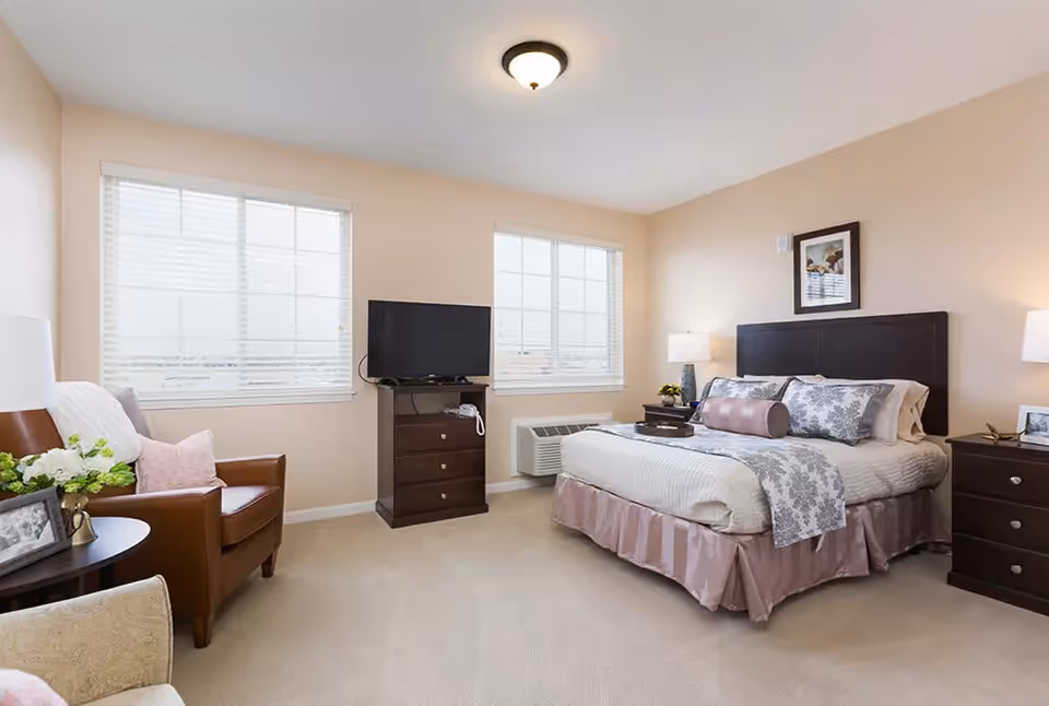 Bright, neatly furnished bedroom with a bed, nightstands, a dresser-mounted TV, and seating by two windows.