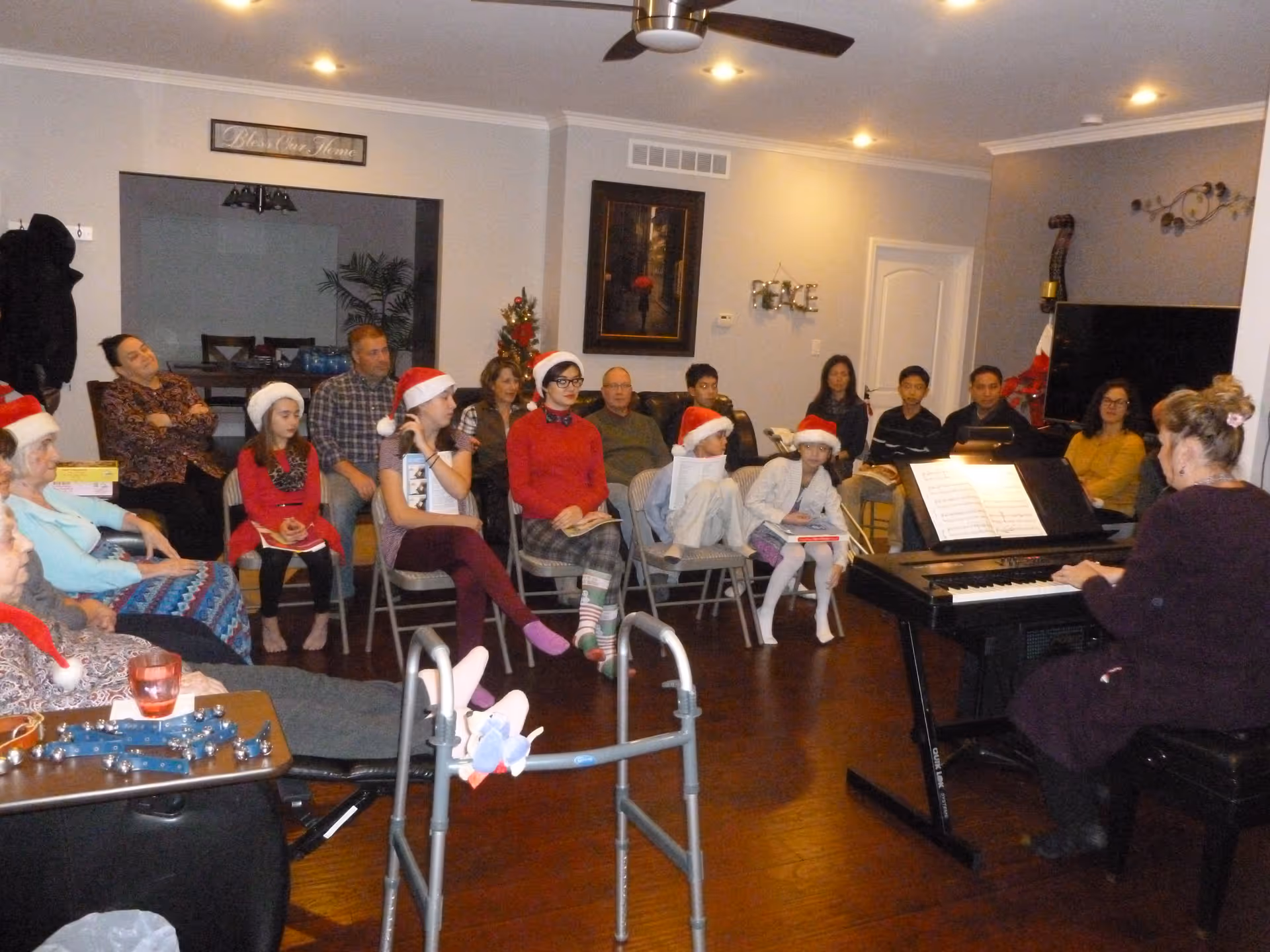A group of people, including children wearing Santa hats, seated in a living room watching a woman play an electric piano. The room is decorated with a small Christmas tree and holiday decorations, and there is a walker in the foreground.