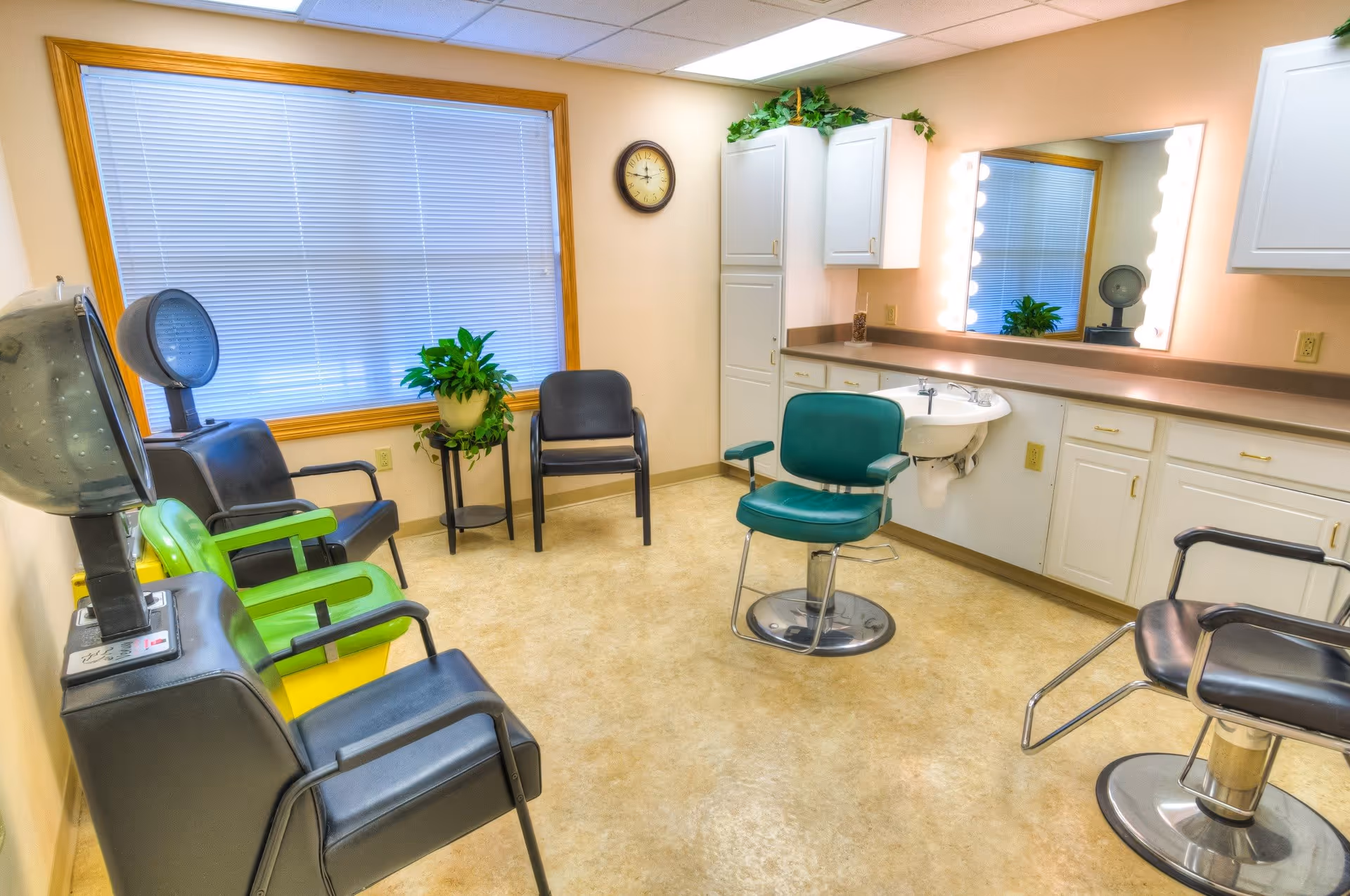 A bright and clean salon room with several chairs including a green salon chair and black waiting chairs. There is a large mirror with lights around it above a countertop with cabinets and a sink. A window with closed blinds and a clock on the wall are also visible, along with a potted plant on a stand.