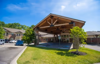 Entrance of The Lodge at Natchez Trace with a large covered driveway supported by stone pillars, surrounded by green grass, trees, and parked cars under a clear blue sky.