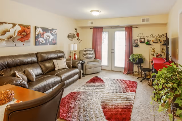 A cozy living room with a dark brown leather sofa, a gray armchair, and a colorful red, white, and gray patterned rug on a carpeted floor. The room features two large glass doors with red curtains, wall art including floral paintings and decorative signs, a small table with a lamp, and several green plants. A TV is visible on the right side of the room.