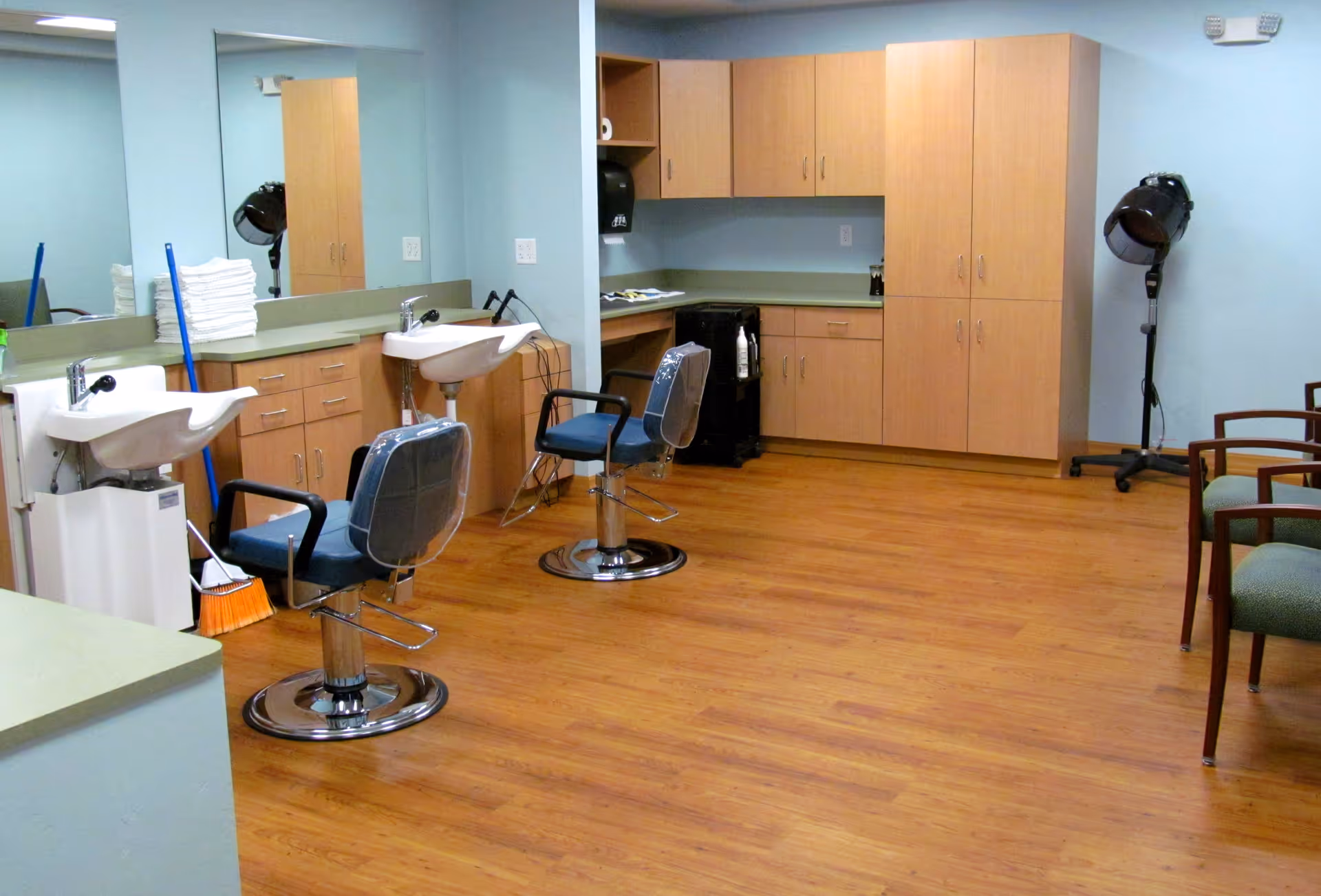 Interior view of a salon area in a senior living facility with two salon chairs facing sinks for hair washing, wooden cabinets and countertops, a hair dryer on a stand, and a few green cushioned chairs along the right wall. The floor is wooden and the walls are painted light blue.