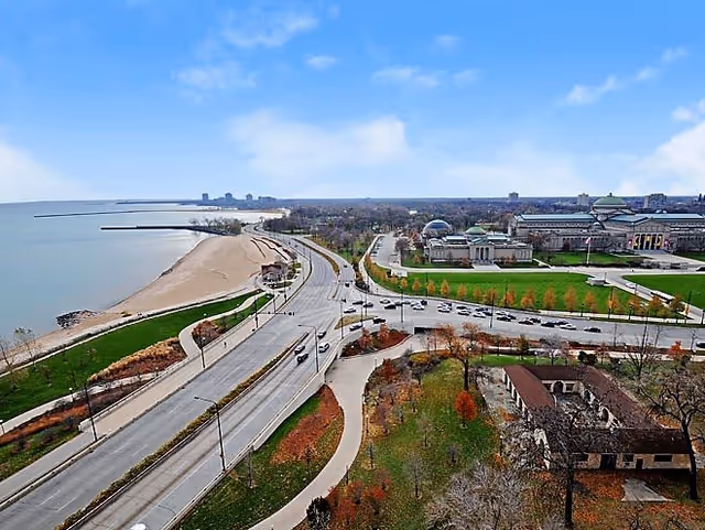 Aerial view of a coastal city area showing a beach along a large body of water, a multi-lane road with cars, green spaces with trees, and several large buildings including a domed structure under a partly cloudy sky.