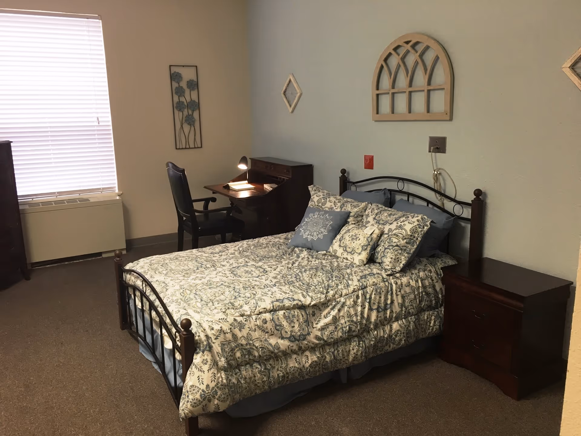 A bedroom with a bed covered in a patterned comforter and multiple pillows. There is a wooden nightstand next to the bed and a wooden desk with a chair and a desk lamp in the corner. The walls are decorated with three pieces of wall art, and a window with closed blinds is visible.