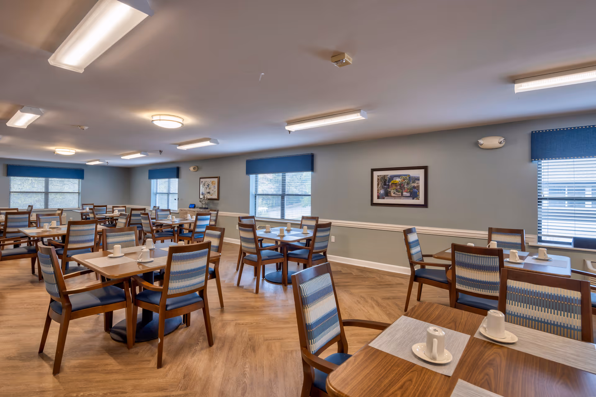 A spacious dining room with multiple wooden tables and chairs arranged neatly. Each table has white cups and saucers placed on light-colored placemats. The room has large windows with blue valances, light blue walls, framed artwork, and wood-patterned flooring. Ceiling lights provide bright illumination.