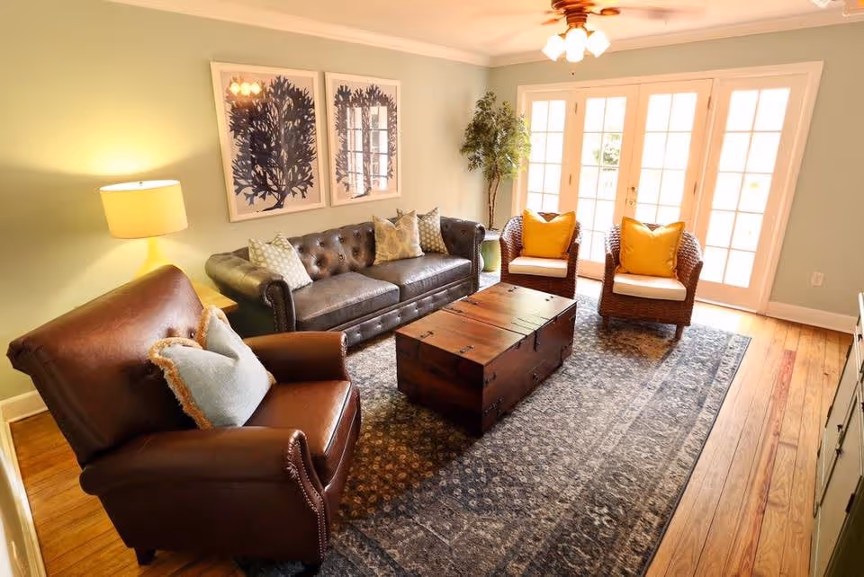 A cozy living room with a brown leather armchair, a dark leather sofa with patterned pillows, two wicker chairs with yellow cushions, a wooden chest coffee table, a patterned rug, a floor lamp, two framed botanical prints on the wall, a potted plant, and French doors letting in natural light.