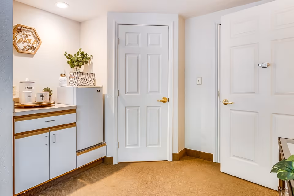 Interior view of a small kitchen area with white cabinets and a mini refrigerator. There are decorative items on the countertop including a tray with containers labeled 'COOKIE' and 'TEA', a small plant, and a basket labeled 'PANTRY'. Two white doors with gold handles are visible, along with beige carpet flooring and white walls.
