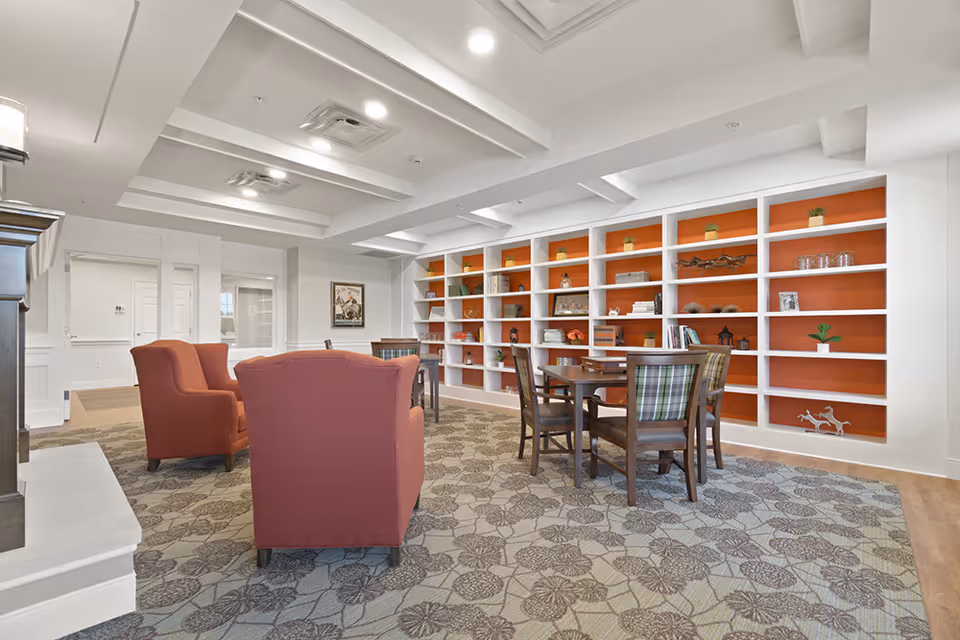 A bright and spacious senior living common area featuring two red armchairs, a round wooden table with four chairs, and a large built-in bookshelf with orange backing filled with decorative items and plants. The room has a patterned carpet, white walls, and a coffered ceiling with recessed lighting.