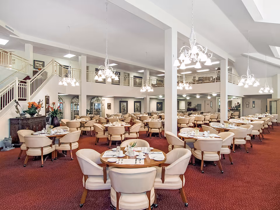 Spacious dining room in a senior living facility with multiple round tables set with white cups, plates, and silverware. The room features beige cushioned chairs, red carpet flooring, white walls, and several elegant chandeliers hanging from the ceiling. There is a staircase leading to an upper level with railings and framed artwork on the walls.