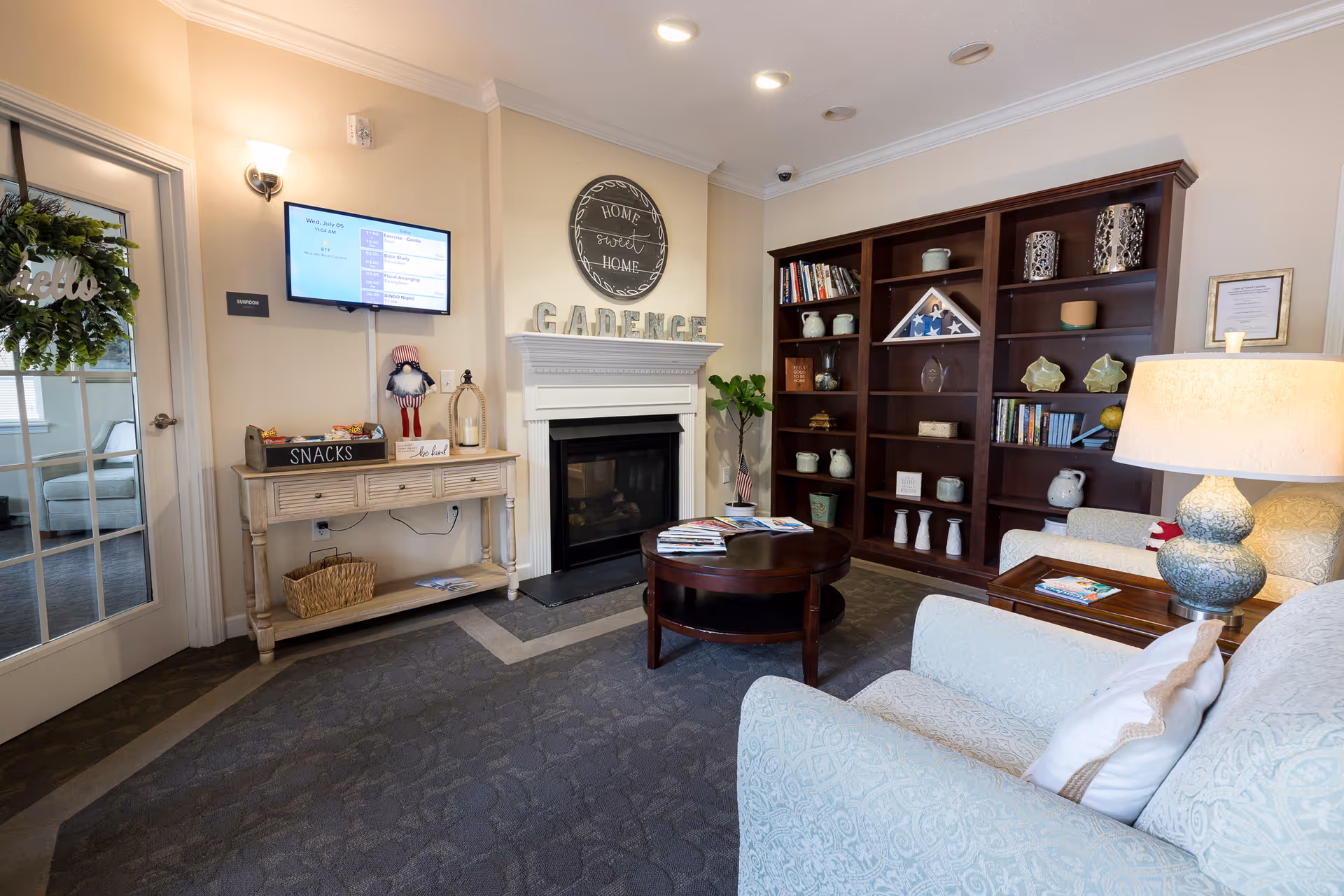 A cozy senior living common area with a fireplace, a large wooden bookshelf filled with decorative items and books, a round wooden coffee table with magazines, and comfortable upholstered armchairs. A small console table holds a snacks box and decorative items beneath a wall-mounted TV screen displaying a schedule. The room has soft lighting and a welcoming atmosphere.