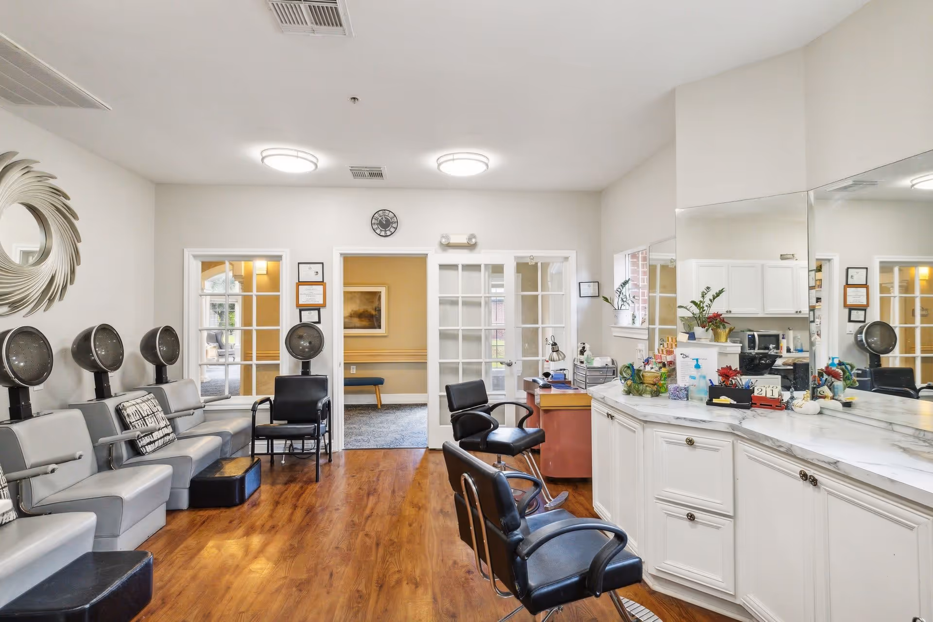 Bright interior salon area with multiple hooded hair dryers and seating on the left, styling chairs and a long mirrored counter on the right.