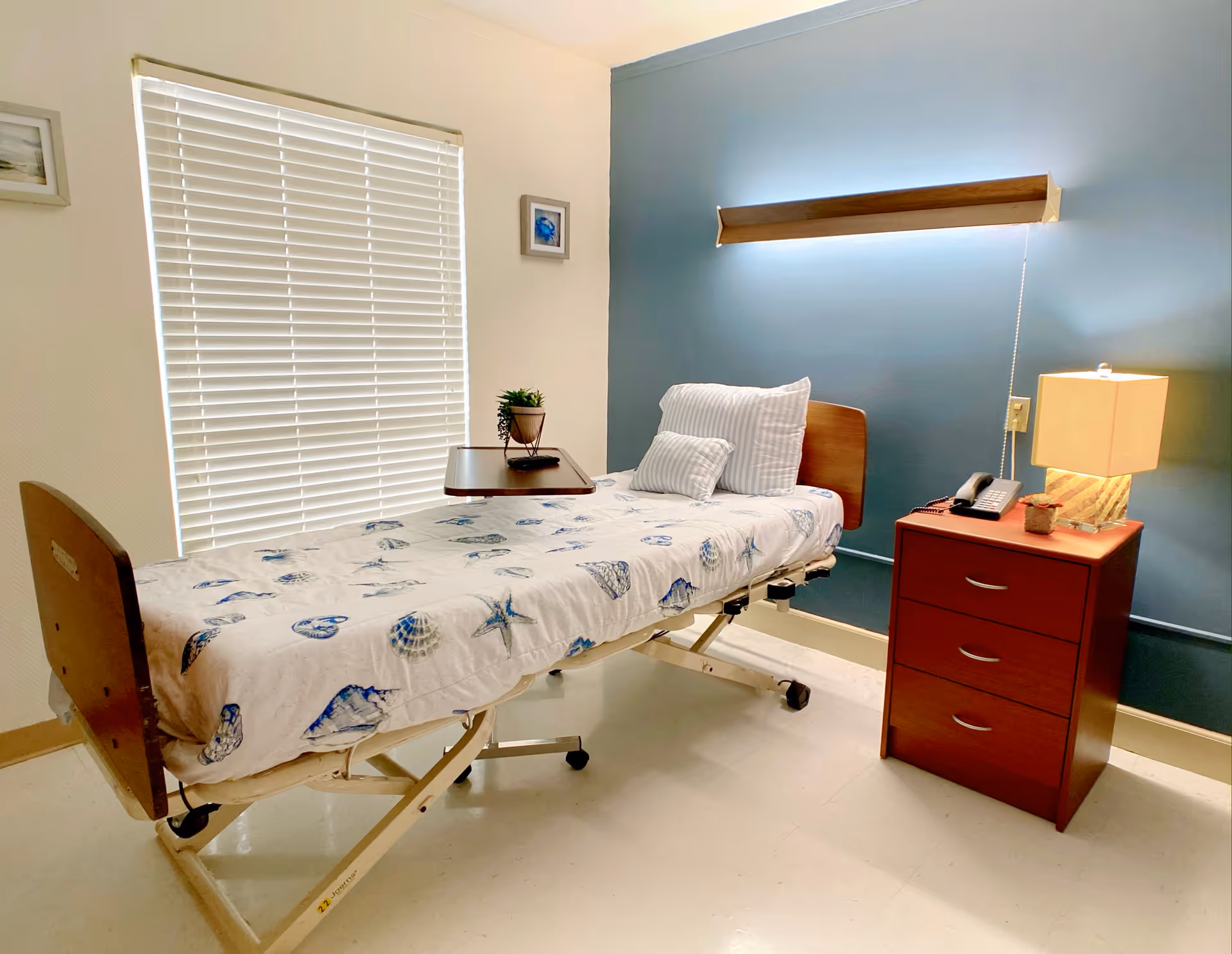A small, neatly arranged room with a hospital-style bed covered with a seashell-patterned blanket and two pillows. Next to the bed is a wooden nightstand with three drawers, a lamp, a telephone, and a small plant. The wall behind the bed is painted blue with a long light fixture mounted above the bed. A window with closed blinds and two framed pictures are on the adjacent wall.
