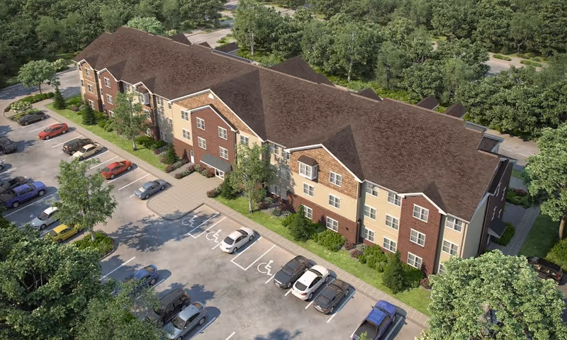 Aerial view of a three-story senior living facility building with a brown roof and a mix of brick and beige siding. The building is surrounded by trees and landscaping, with a parking lot in front that includes several cars and designated handicap parking spaces.