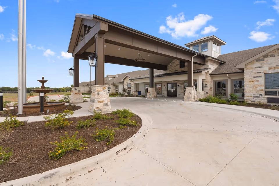 Front entrance of a senior living facility with a large covered porte-cochere, stone facade, driveway, and fountain.