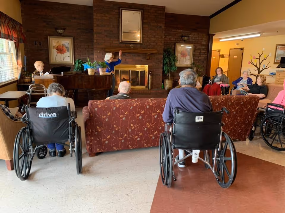 A group of elderly people, some in wheelchairs, seated in a common living area with a brick fireplace and a piano. One person is standing near the fireplace, possibly leading an activity or speaking to the group. The room has warm lighting and framed floral artwork on the walls.