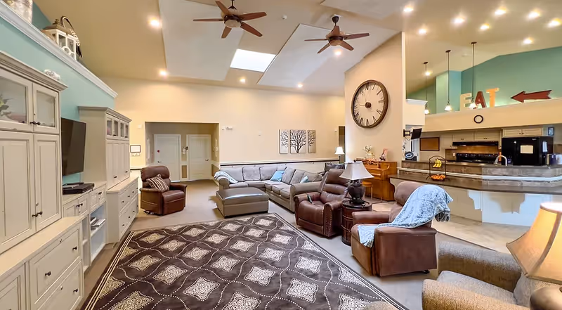 Spacious assisted-living common room featuring a sectional sofa and leather recliners around a patterned rug, TV cabinets, ceiling fans, and an open kitchen/bar area.