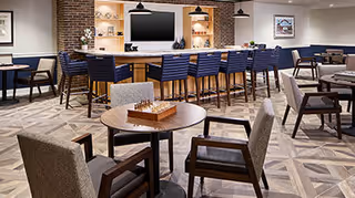 A spacious and modern common area with a large bar counter featuring high chairs, a wall-mounted TV, and shelves with decorative items. Several tables and comfortable armchairs are arranged throughout the room, with a chessboard on one of the tables. The floor has a patterned design, and the lighting includes pendant lamps hanging from the ceiling.