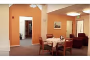 Interior view of a senior living facility common area with a small dining table covered with a white tablecloth and four wooden chairs. In the background, there are two armchairs and a hallway leading to other rooms. The walls are painted in a warm beige color and there is a framed picture hanging on the wall.