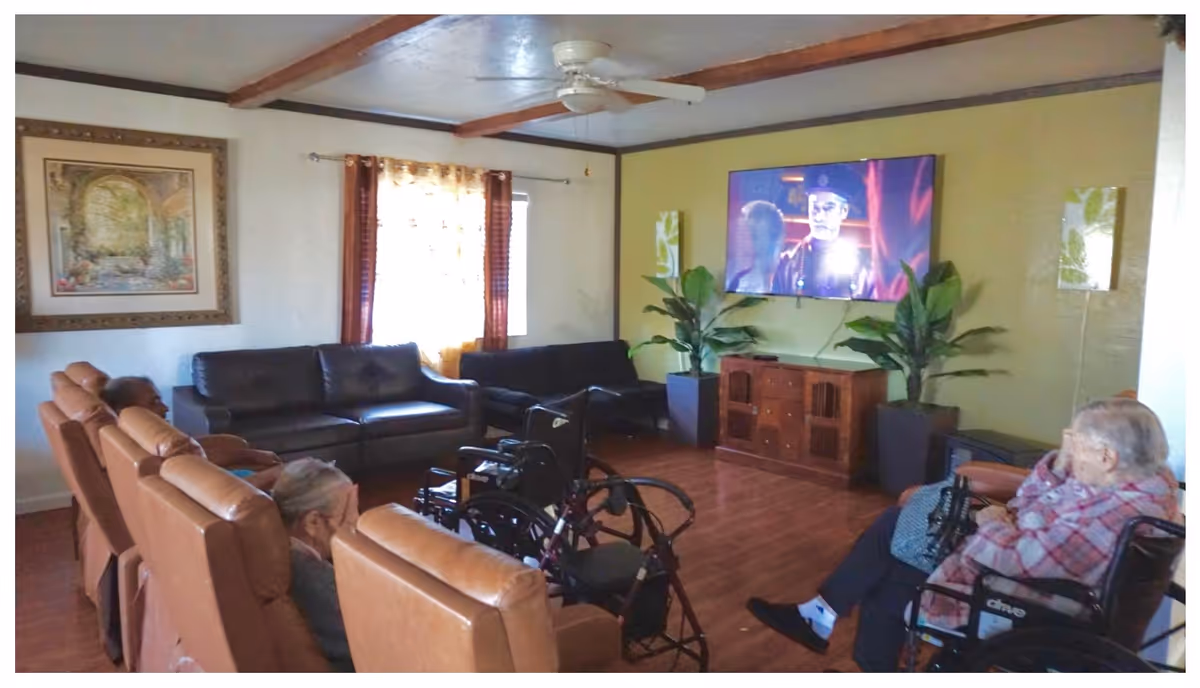 A cozy living room in a senior living facility with several elderly residents seated in comfortable chairs and wheelchairs, watching a television mounted on a green wall. The room has wooden flooring, a ceiling fan, two large potted plants flanking a wooden cabinet under the TV, and a framed painting on the wall near a window with curtains.
