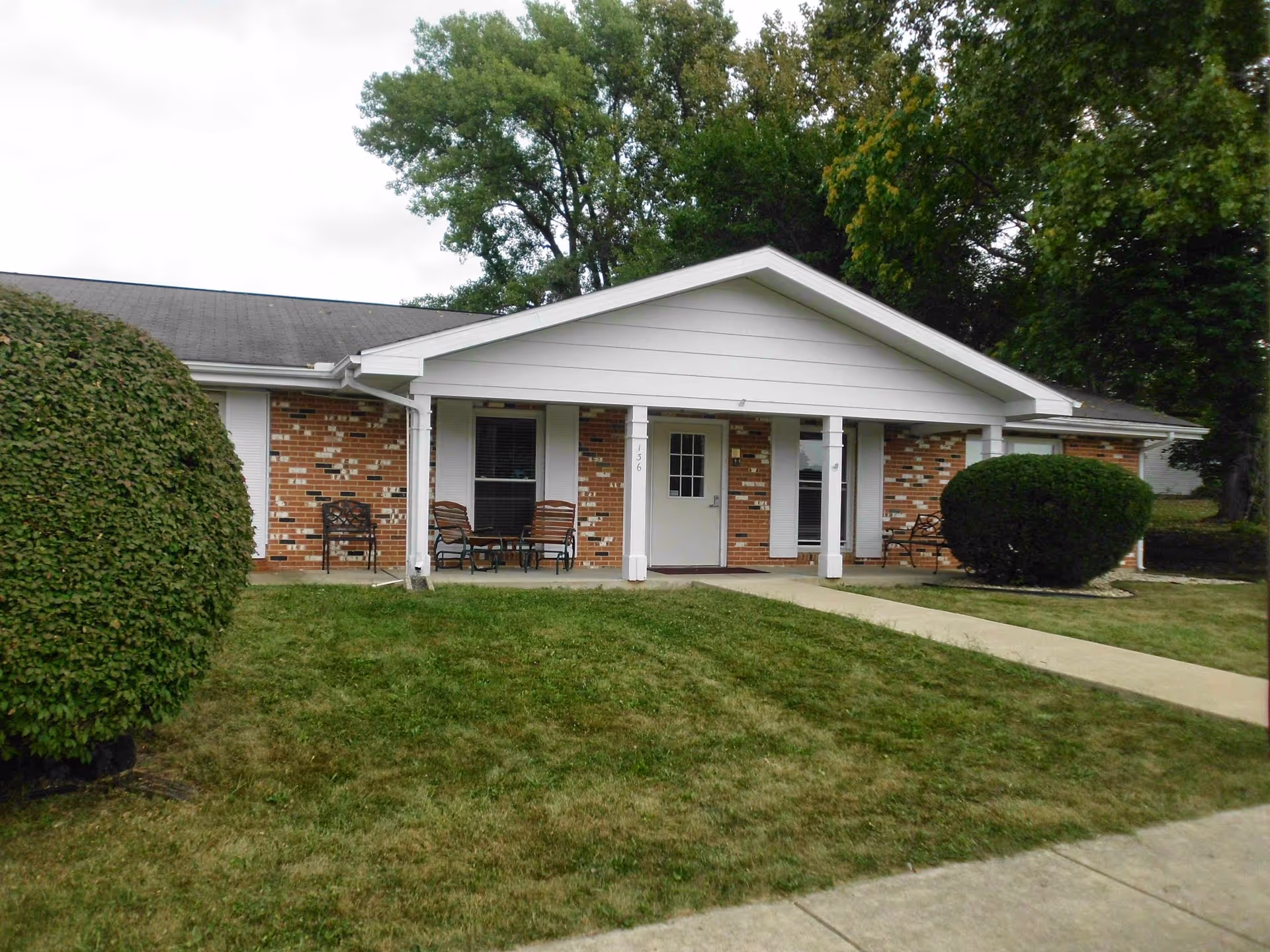 Single-story brick building with a covered front porch, chairs, walkway, and trimmed bushes on a lawn.