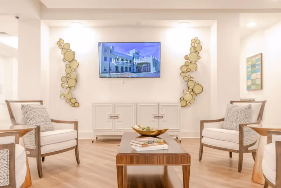 A bright and modern living room area with four white cushioned armchairs arranged around a wooden coffee table. The coffee table has a decorative bowl and some books on it. On the wall, there is a flat-screen TV displaying an image of a building with the text 'Welcome Home'. Two decorative gold circular wall art pieces flank the TV. The room has light wood flooring and white walls.