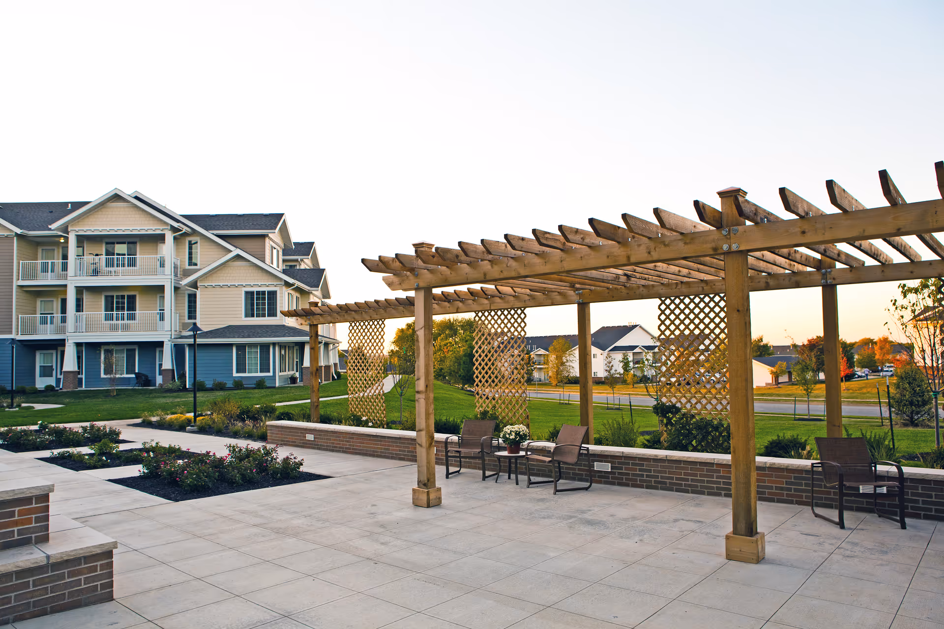 Outdoor patio with a wooden pergola, chairs, planters, and a grassy courtyard in front of a multi-story residential building.