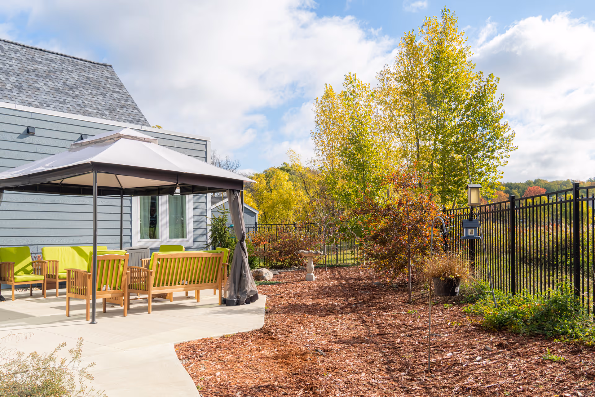 Outdoor patio with a canopy and green-cushioned wooden seating next to a landscaped yard and metal fence under a partly cloudy sky.