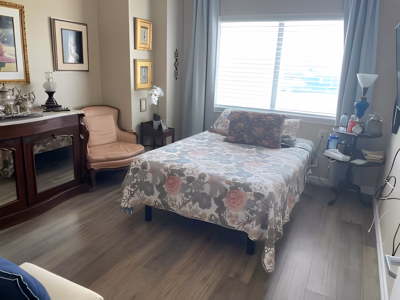 A cozy bedroom with a bed covered in a floral patterned bedspread, a large window with blinds and curtains, a vintage armchair, a wooden side table with a lamp and books, and framed pictures on the walls.