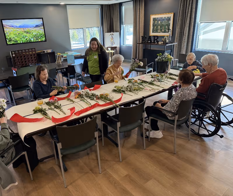A group of elderly women seated around a long table in a well-lit room, engaging in a flower arranging activity with various flowers and red ribbons spread out on the table. A caregiver stands nearby assisting them. The room has large windows, a television mounted on the wall, and a small bar area in the background.