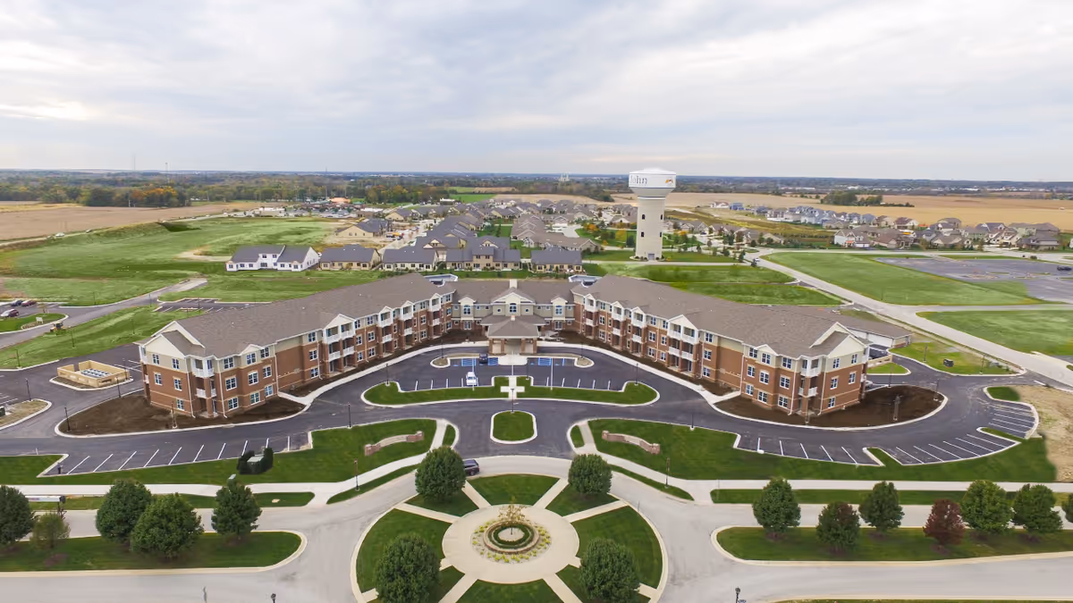 Aerial view of a curved three-story senior living building with a circular driveway, parking lots, lawns, and surrounding neighborhood.