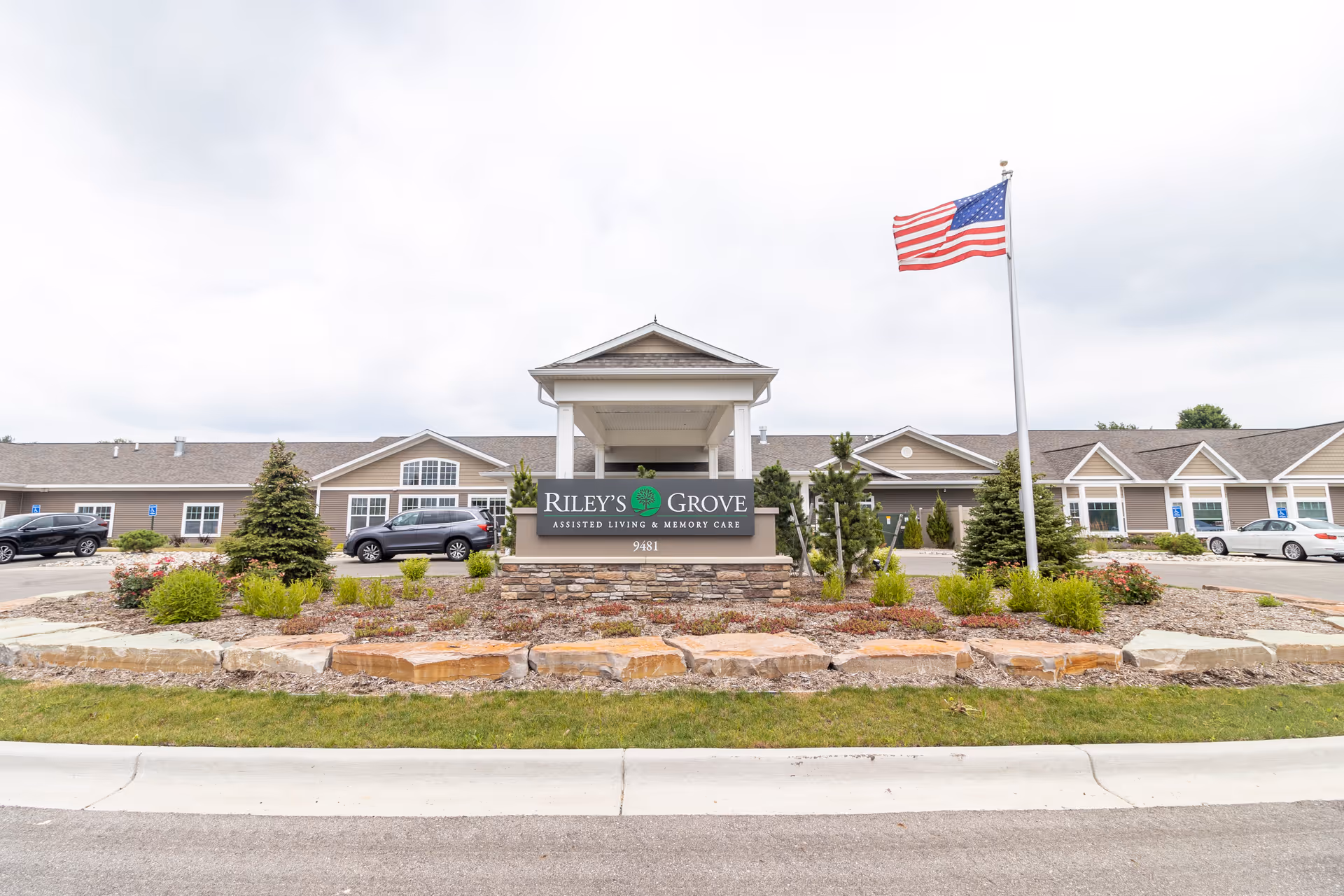 Front exterior view of Riley's Grove Assisted Living & Memory Care facility with a landscaped area featuring shrubs and rocks, an American flag on a flagpole, and several parked cars in the parking lot.