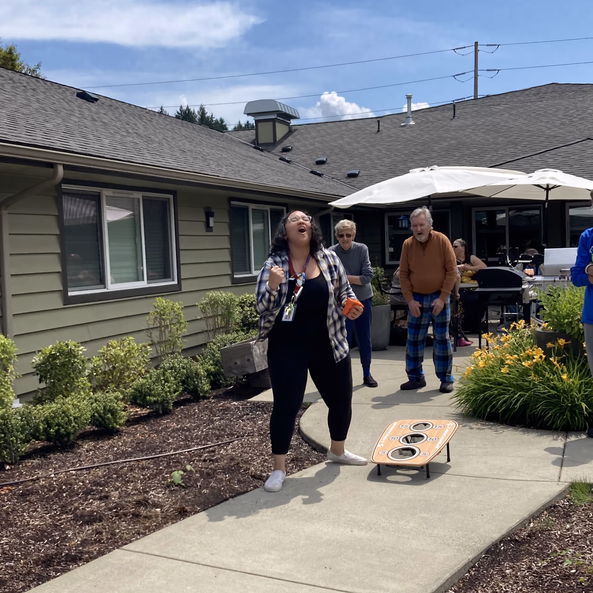 A group of people enjoying an outdoor activity at a senior living facility. A woman in the foreground is enthusiastically playing a bean bag toss game on a paved walkway. Behind her, an older man and woman watch and smile. The setting includes a building with green siding, windows, and a patio area with umbrellas and plants.