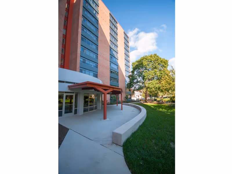Exterior view of a multi-story brick apartment building with large windows, a covered entrance supported by red beams, a curved concrete walkway, and a green lawn with trees under a blue sky with some clouds.