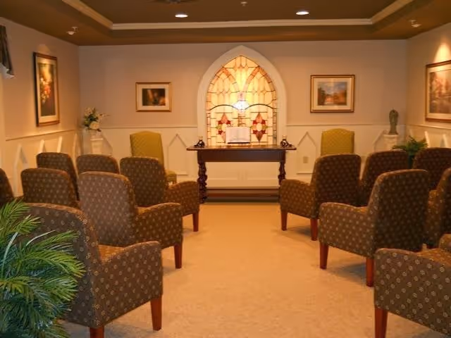 A small chapel or meditation room with rows of upholstered chairs facing a wooden table with an open book and two small lamps. The back wall features a decorative stained glass window, and the room is softly lit with framed artwork and plants on the sides.