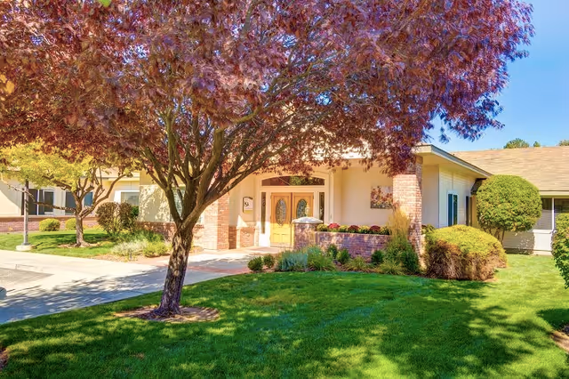 Exterior view of Overland Court Senior Living facility showing a well-maintained lawn, a tree with reddish leaves, and the entrance with double wooden doors under a covered porch.