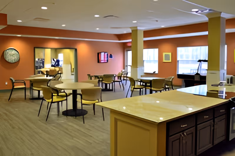 A spacious common area with several round tables and chairs arranged for seating. The walls are painted orange with large windows allowing natural light to enter. There is a kitchen island with a marble countertop in the foreground and a TV mounted on the far wall. The room has a warm and inviting atmosphere.