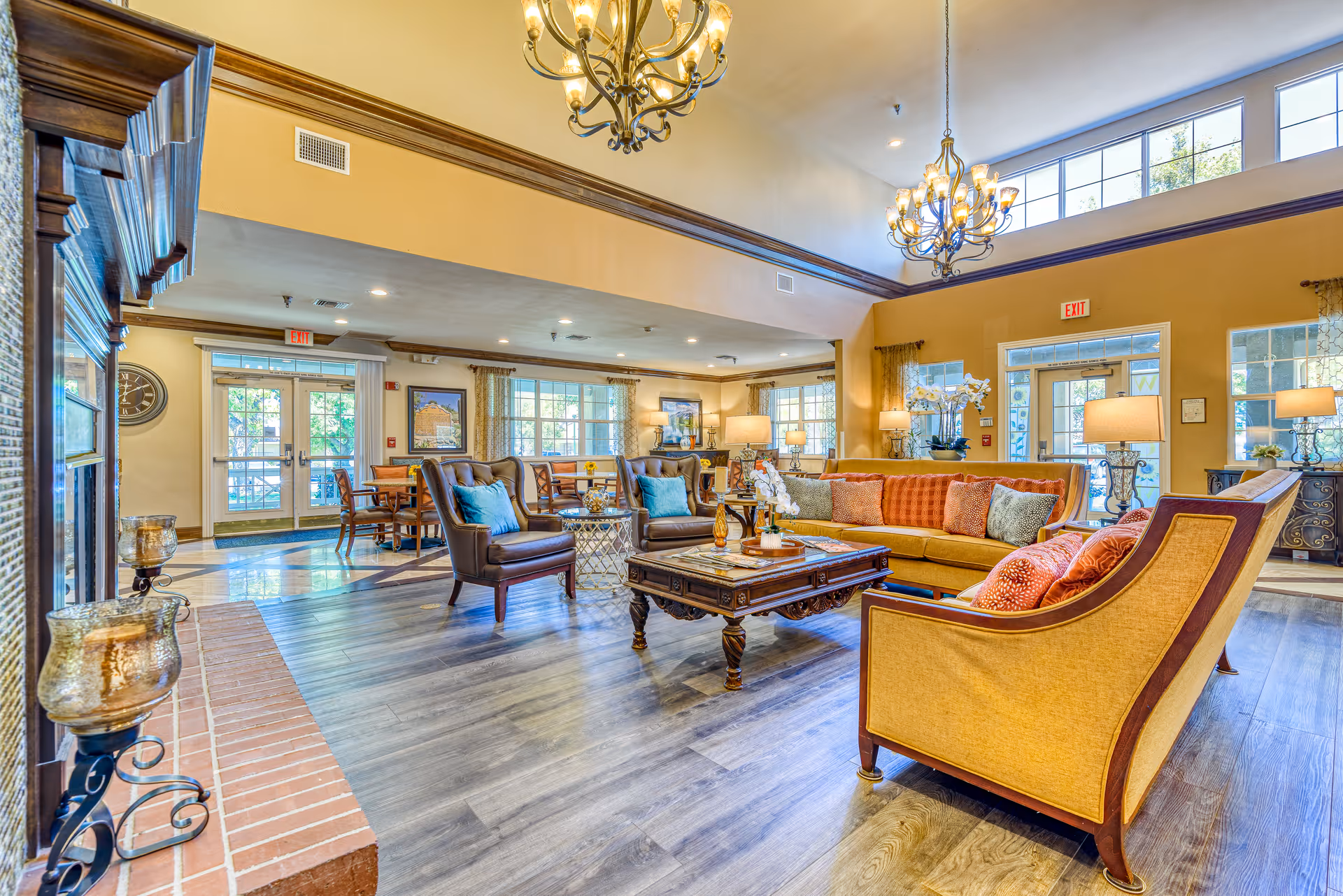 A spacious and well-lit living room area in a senior living facility with high ceilings and large windows. The room features comfortable seating including a yellow sofa with orange and patterned pillows, two brown leather armchairs with blue pillows, and a wooden coffee table with decorative items. There are chandeliers hanging from the ceiling, multiple table lamps, and a brick fireplace on the left side. In the background, there are dining tables and chairs near large windows with curtains, and double glass doors leading outside.