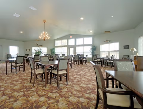 A spacious dining room with multiple wooden tables and cushioned chairs arranged neatly on a patterned carpet. Large windows allow natural light to fill the room, and there are decorative plants and wall-mounted fish sculptures. A chandelier hangs from the ceiling.