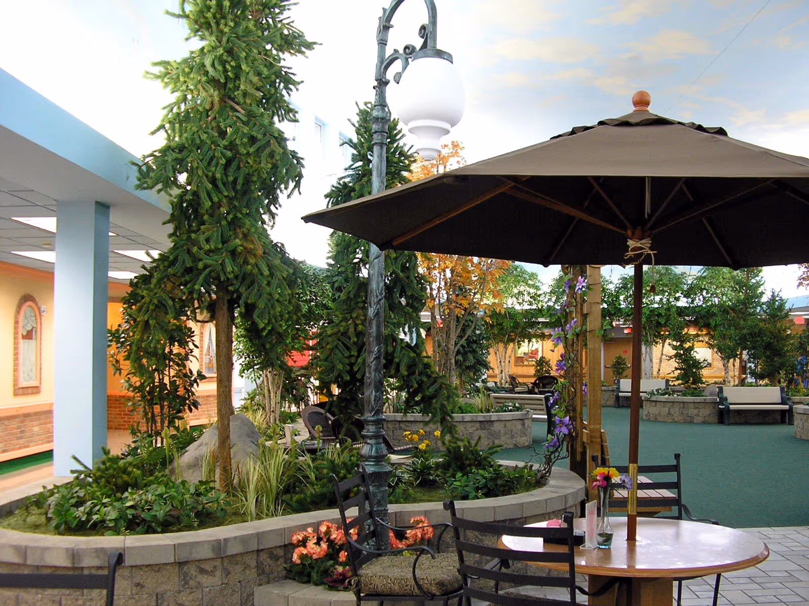Indoor garden area with trees, plants, and flowers surrounded by a low stone wall. There is a round table with a large brown umbrella and chairs around it. In the background, there are benches and more greenery, with a ceiling painted to resemble the sky.