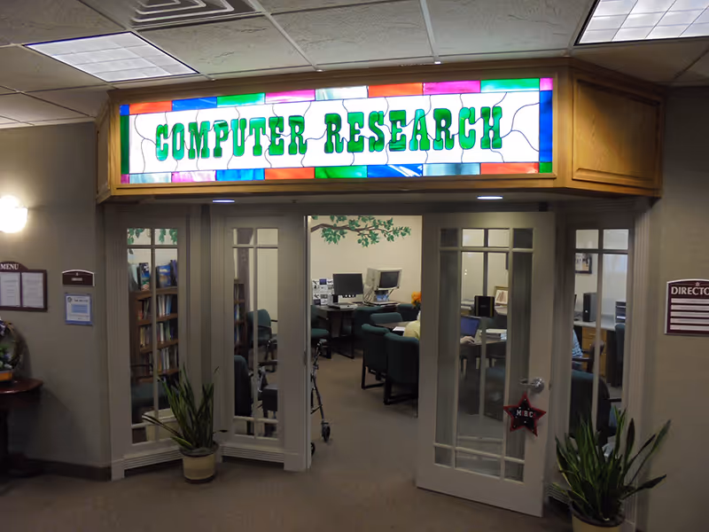 Entrance to a computer research room with double glass doors, a colorful stained glass sign above reading 'COMPUTER RESEARCH', and several computer workstations visible inside. There are potted plants on either side of the entrance and wall-mounted lights illuminating the area.